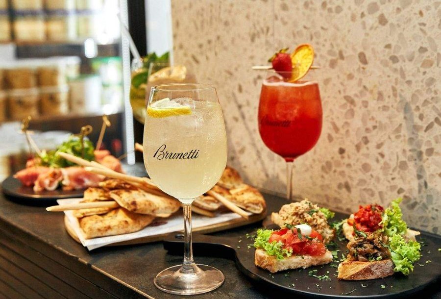 Two cocktails on a bench with plates of food beside them