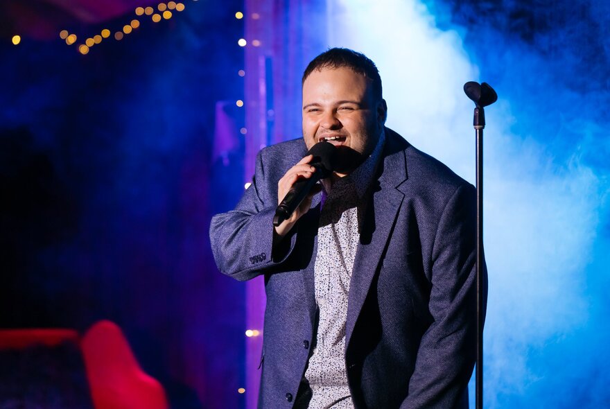 A man in a suit jacked leaning forward slightly on stage with a bit smile and holding a microphone. 
