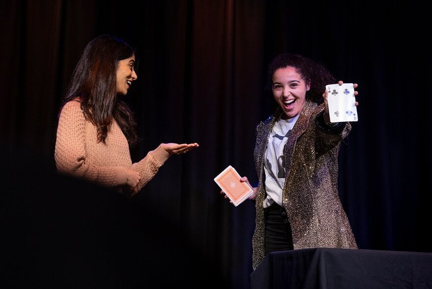 Two teenage performers on a darkened stage, doing tricks with an oversized deck of cards.