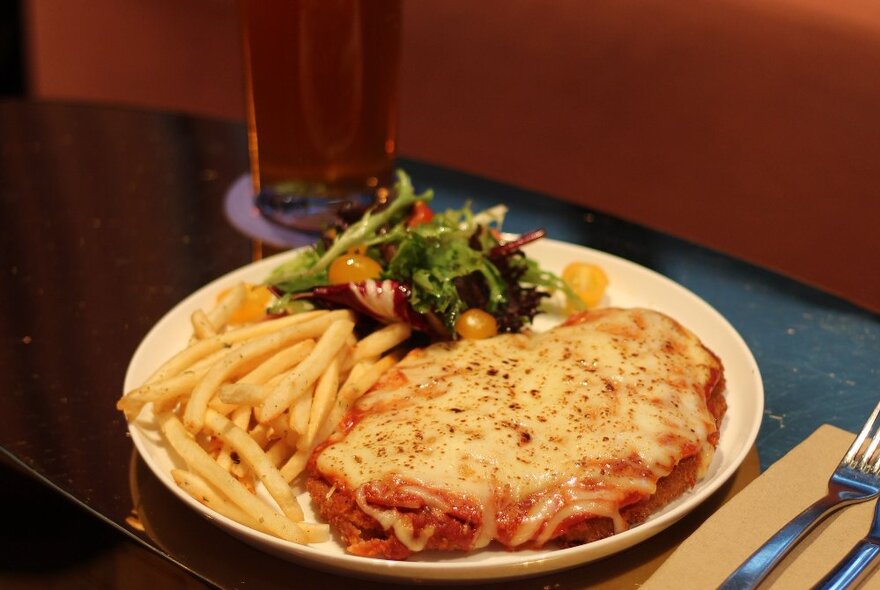 A plate of chicken parma with fries and salad, on a table, a glass of beer in the background.