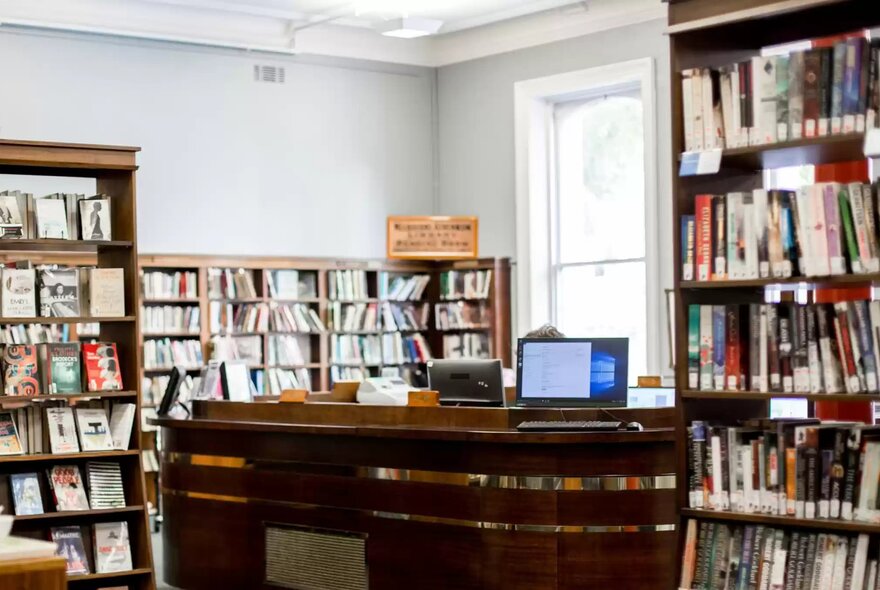 Interior of Melbourne Athenaeum Library featuring a dark wooden service desk with a computer monitor on top, flanked by tall bookshelves filled with books.