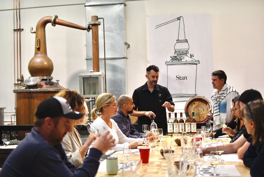 People seated at a table with distilling equipment and multiple tasting glasses and bottles of spirits.