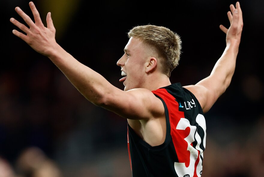 An Essendon AFL player celebrating during a match with his arms raised in the air above his shoulders.