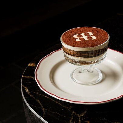 A classic layered tiramisu dessert served in a footed glass bowl with a branded cocoa powder stencil detail, presented on a white plate against a dark table surface.