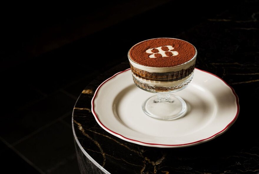 A classic layered tiramisu dessert served in a footed glass bowl with a branded cocoa powder stencil detail, presented on a white plate against a dark table surface.