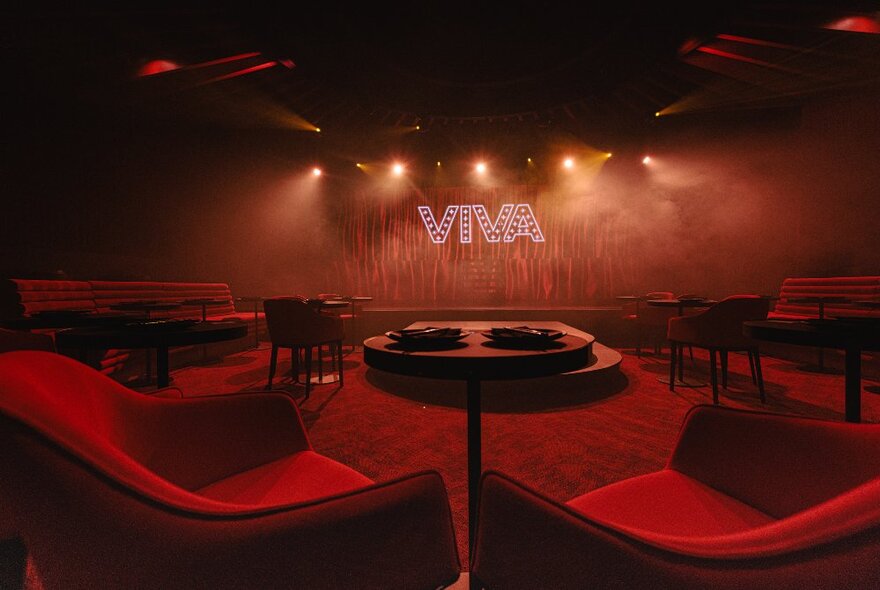 Red smoky interior of Viva, with two club chairs and a small round table between them, with a view of the stage that features the sign VIVA on the stage curtain.