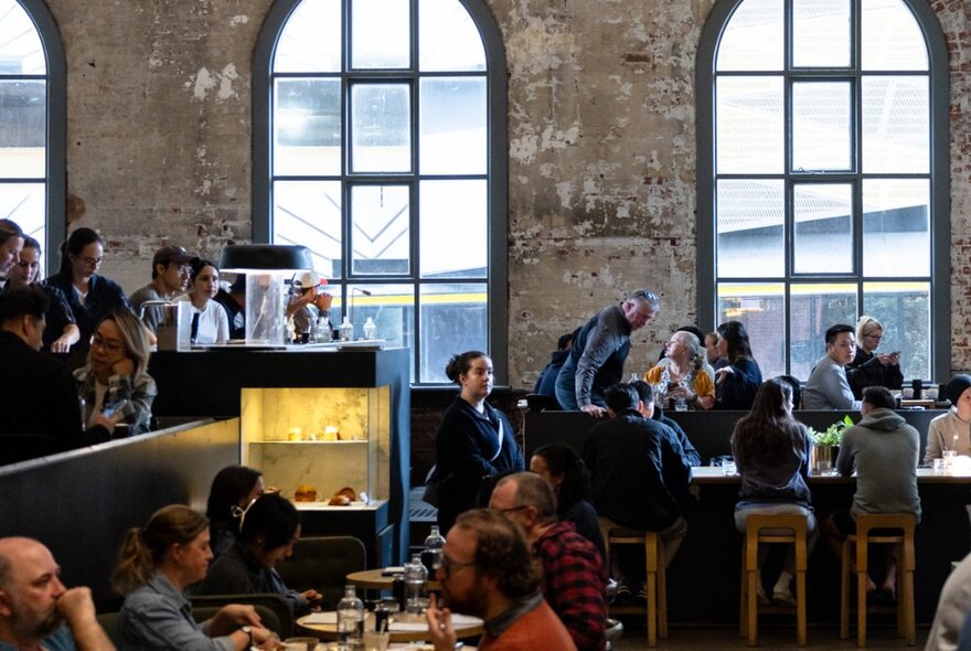Diners seated at communal tables in a soaring space with high arched windows and brick walls.
