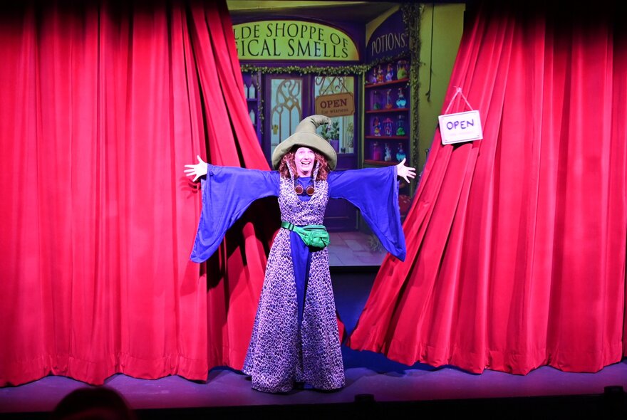 Winni Wizard, a kids comedian, wearing a purple wizard's robe and hat standing on a stage with arms outstretched in front of a slightly parted red curtain.