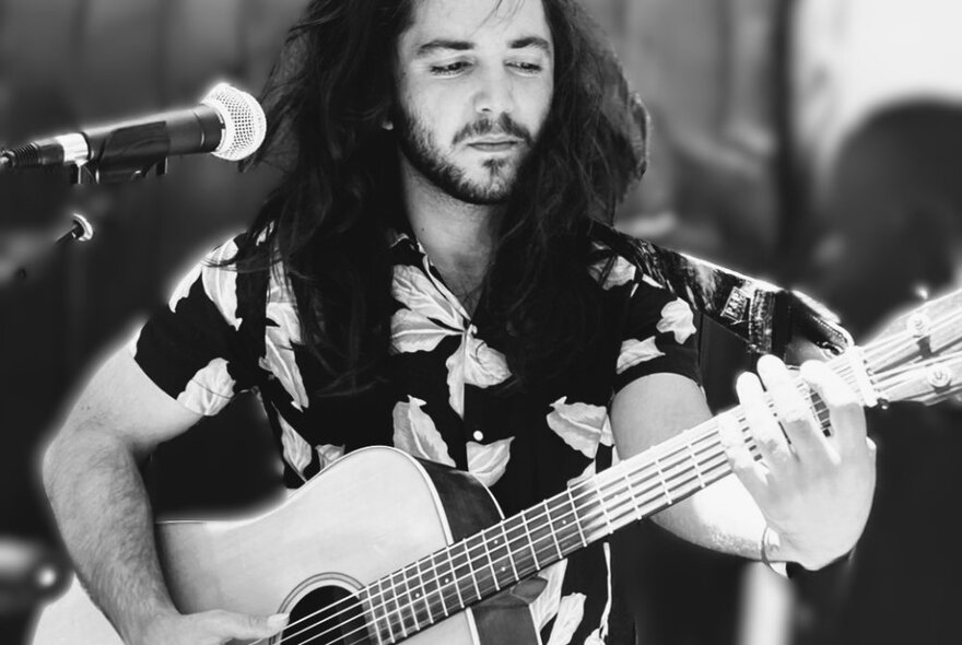 Musician with long dark hair and beard playing guitar at a microphone, wearing a dark patterned shirt.