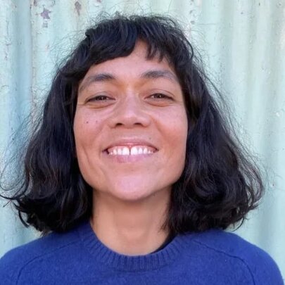 Headshot of a smiling person with medium-length, dark, wavy hair, wearing a blue sweater and standing in front of a light blue corrugated metal wall.