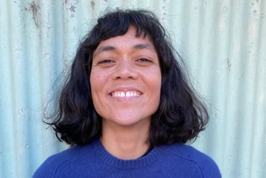 Headshot of a smiling person with medium-length, dark, wavy hair, wearing a blue sweater and standing in front of a light blue corrugated metal wall.
