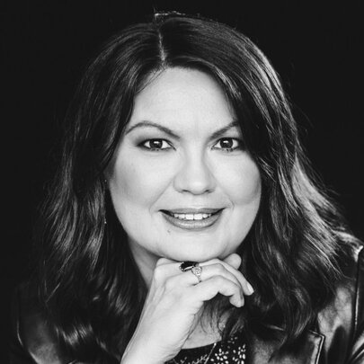A black and white headshot of author Katya de Becerra, smiling while resting her chin on her right hand.