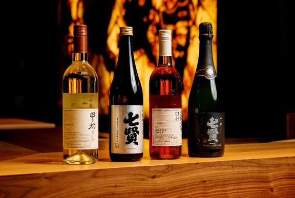 Two bottles of Japanese sake and two bottles of wine lined up on a wooden shelf, against a fire-like background.