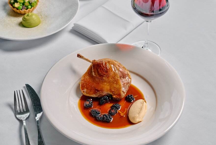 A white plate of carefully arranged food at a fine dining restaurant with silver cutlery, cloth napkins and a glass of red wine.