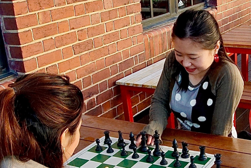 Two women playing chess at an outdoor table next to a brick wall.