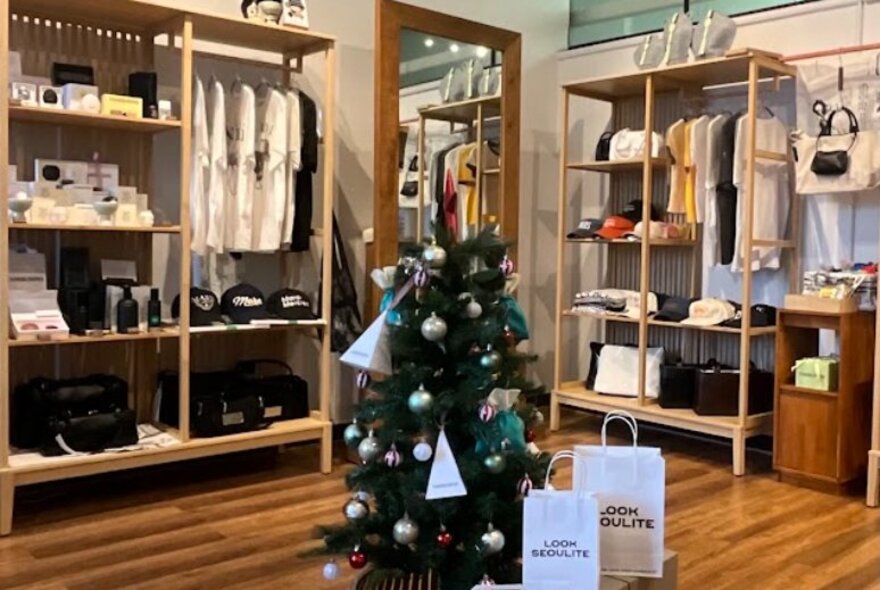 Small store with a central Christmas tree surrounded by shopping bags, the walls lined with display shelves stocked with clothing and ornaments.