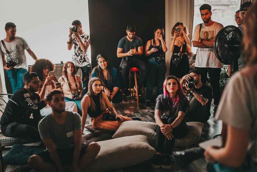 A group of people gathered informally indoors, some sitting on the floor, others standing, listening to a speaker.