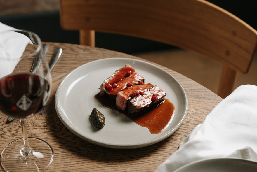 Looking across at a plate with meat and a vivid red sauce, with a glass of red wine next to the plate. 