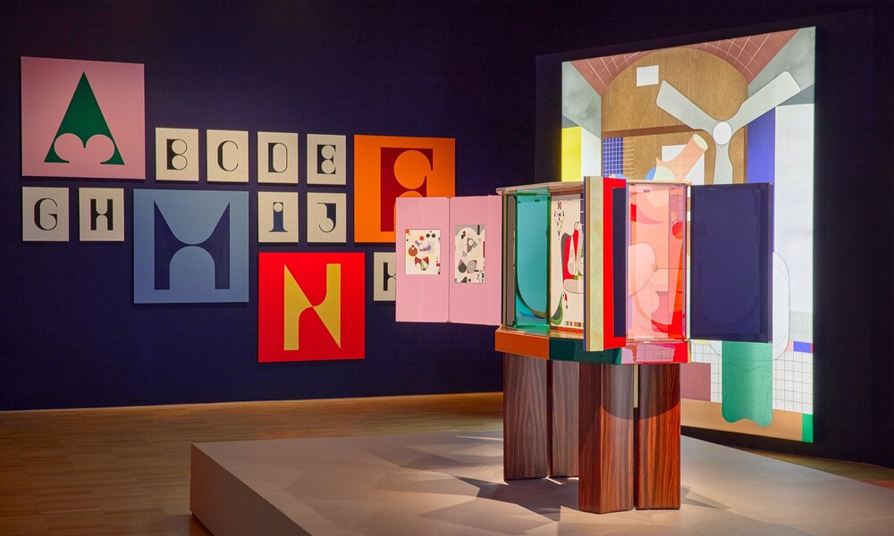 A colourful art exhibition with a sculpture and letter painting on the walls.