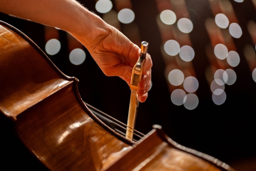 A hand holding a bow and playing a cello, abstract white circular lights in the background.