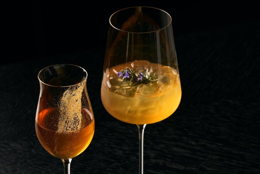 Two orange coloured drinks in wine glasses against a black background.