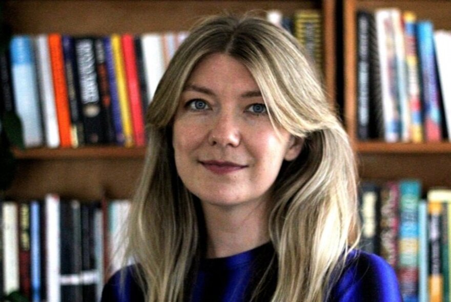 Portrait of Jo Hall, with a slightly smiling expression, standing in front of a wooden bookshelf lined with colourful books.
