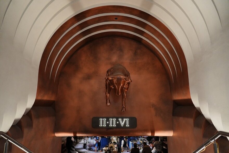 An underground archway entrance to a basement restaurant with a copper-toned background featuring a bull sculpture and the Roman numerals II-II-VI.
