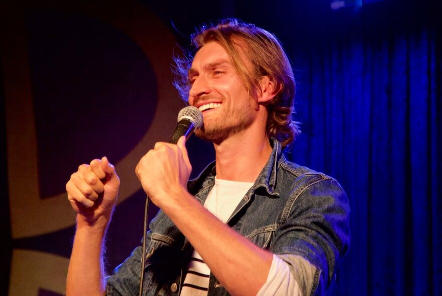 A man on stage with a microphone, wearing a denim shirt with a white striped t-shirt underneath.