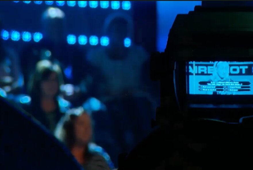 The glowing back of a digital camera, seen with a studio audience sitting in a darkened theatre behind it.