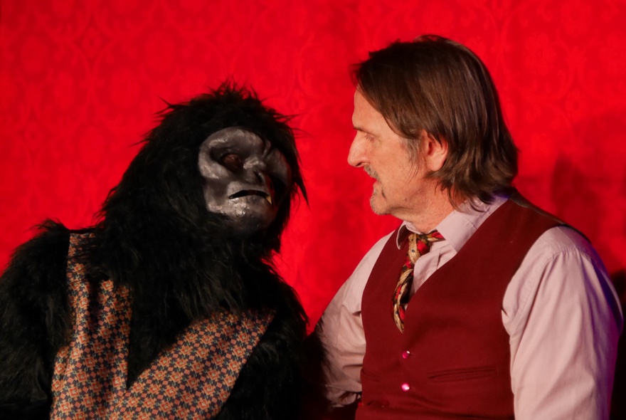 An actor on stage facing a person in a gorilla suit who is wearing a waistcoat.
