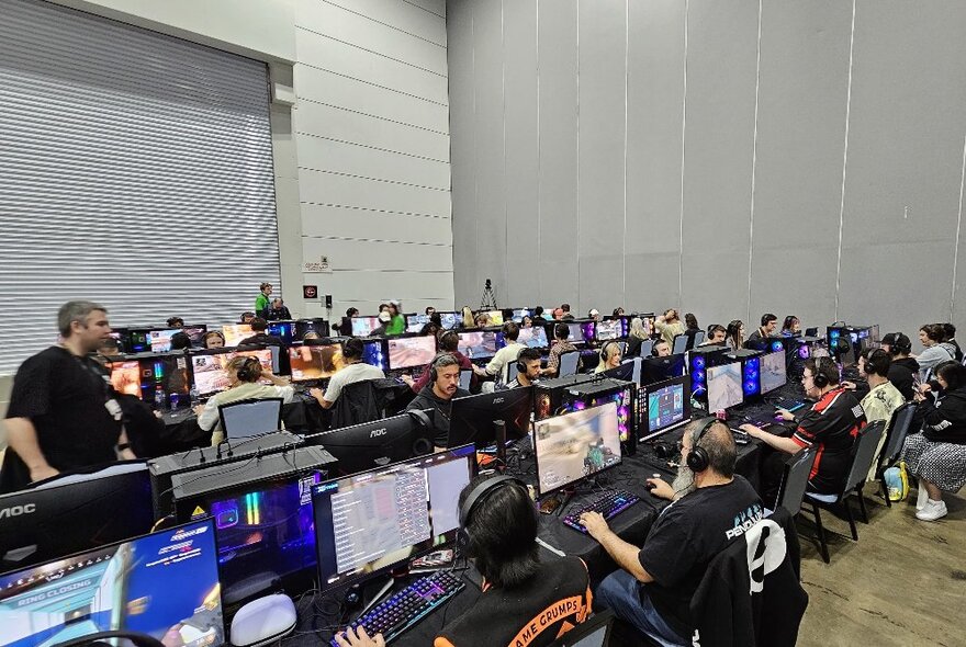 Rows of people at PC gaming consoles playing video games inside a large expo hall.