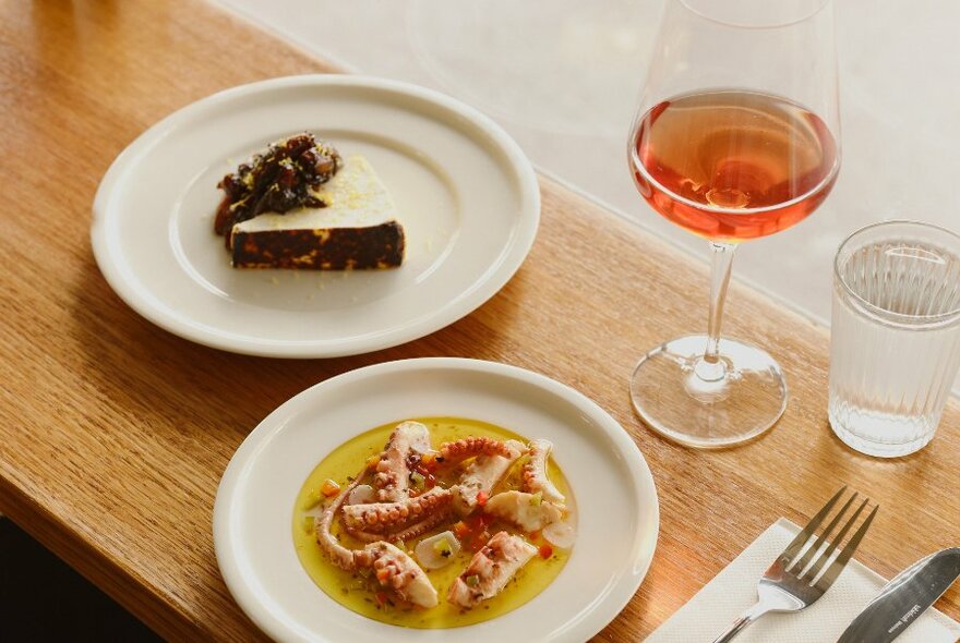 Two white plates of food, a glass of wine and a glass of water, on a wooden table top.