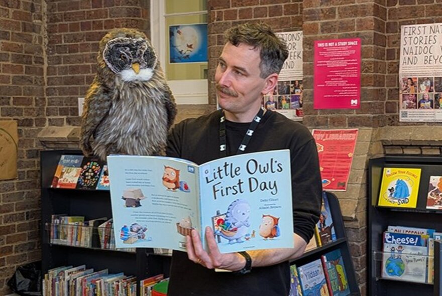 A library staff member holding a large owl puppet in one hand and a picture book in his other hand, standing in the children's section of a library.