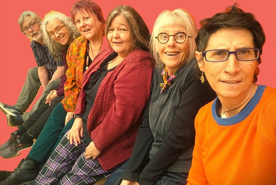 Six older comedians seated in a row looking to their left.