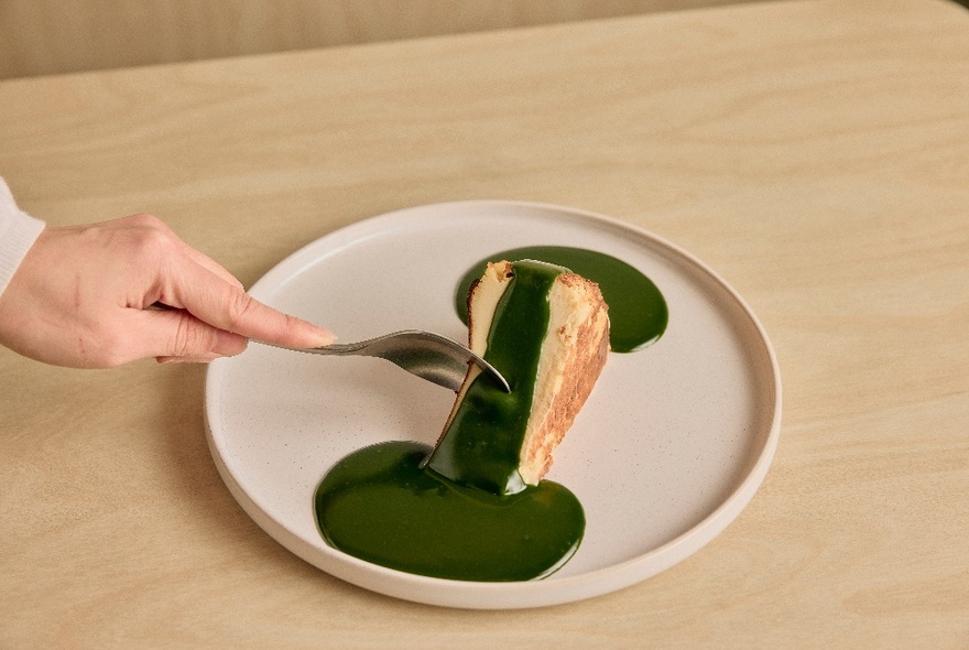 A hand using a spoon to break into a slice of cake covered in a green match sauce.