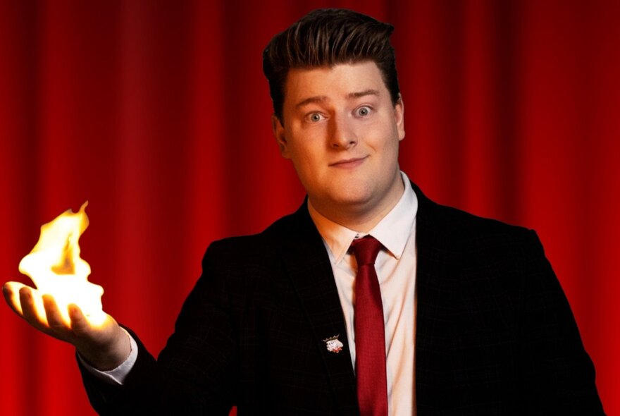 Comic magician wearing a suit and tie, his outstretched hand on fire.