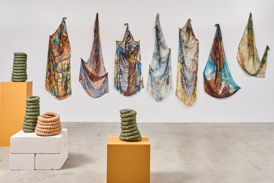 Textiles hanging on a white wall, and woven baskets on plinth displays in the white gallery space at Craft Victoria.