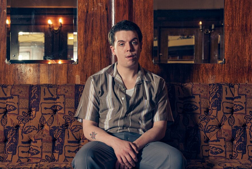 Comedian Ed Night, sitting on a cushioned bench seat in a bar, looking somewhat forlorn.
