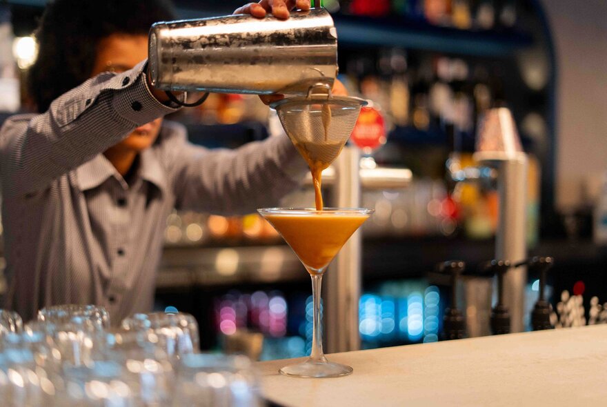 A bartender straining a cocktain into a glass at a bar. 