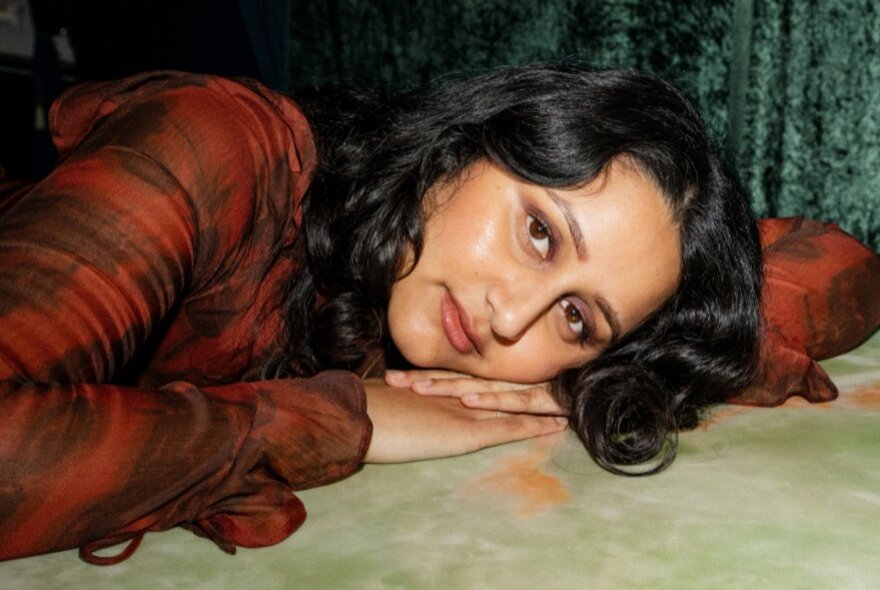 A young woman with wavy, dark hair is resting her head on her hands while lying down, she wears a dark orange-red long sleeve top.