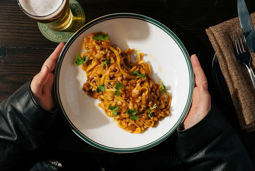 Overhead view of a pair of hands holding a white plate of pasta, a glass of beer also in the frame.