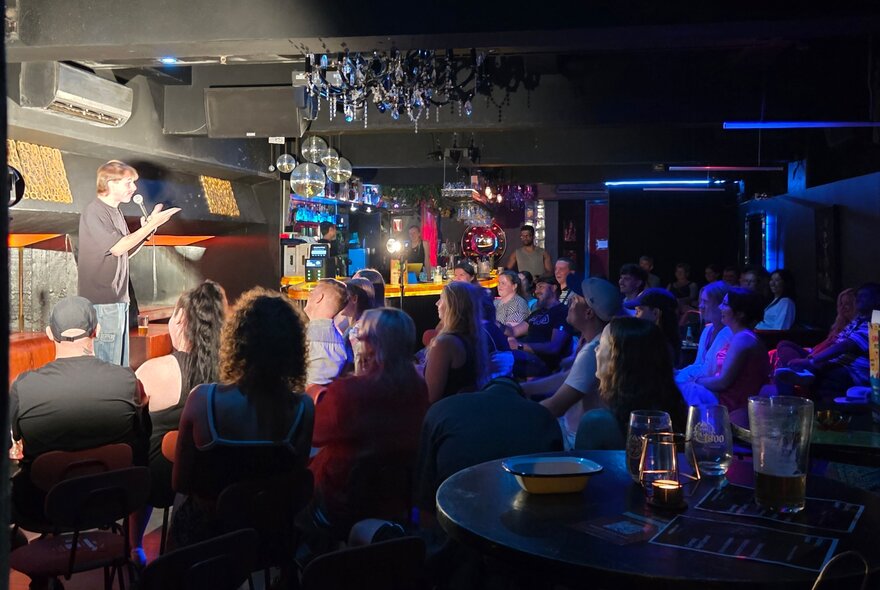 People seated in a dark bar venue watching a performer illuminated on stage, standing at a microphone.