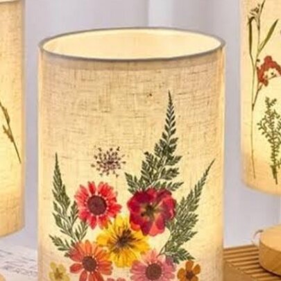 Three illuminated lamps with cylindrical parchment or fabric shades, decorated with various pressed flowers and small human figures. 