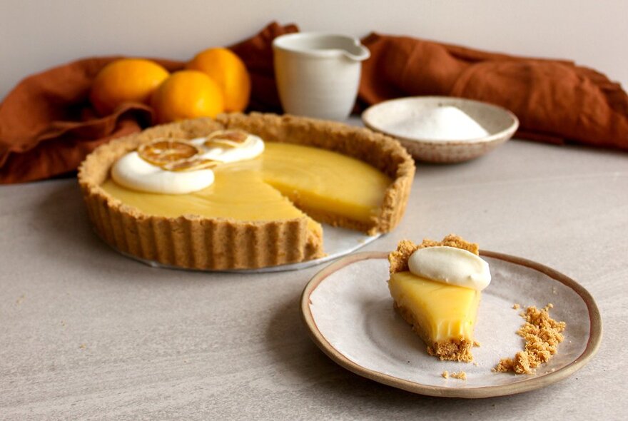 A slice of lemon tart on a plate, the rest of the lemon tart behind it, with ceramics and lemons in the background.