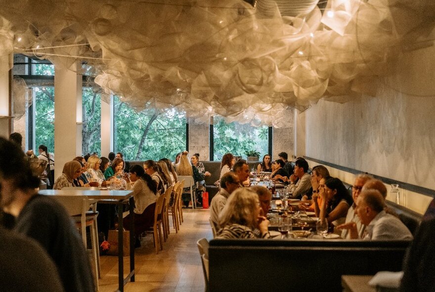 Restaurant interior with people seated at tables along a wall and against large windows, suspended white mesh on ceiling diffusing the lighting.