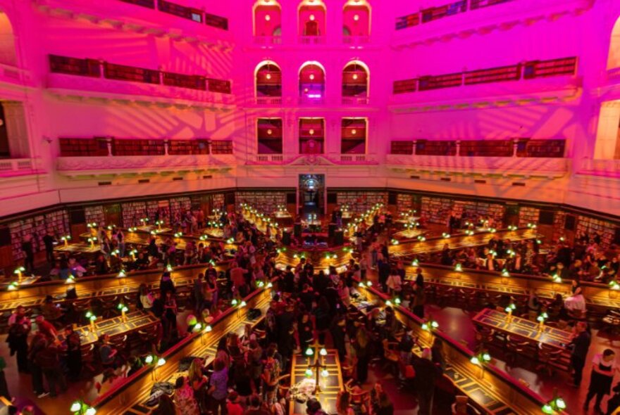 The Dome reading room of State Library Victoria showing the upper level balconies, and rows of desk fanning out from the centre, all bathed in a pink light.