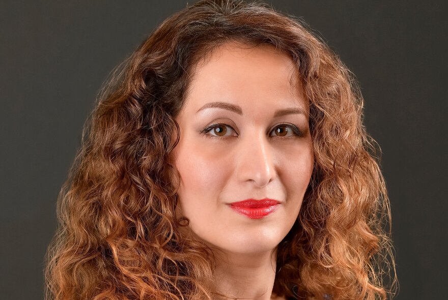 Woman with long curly brown hair, parted on the side, wearing red lipstick.