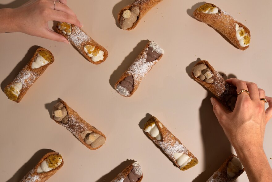 Looking down on multiple cannoli arranged in a pattern with two different hands reaching to grab them. 