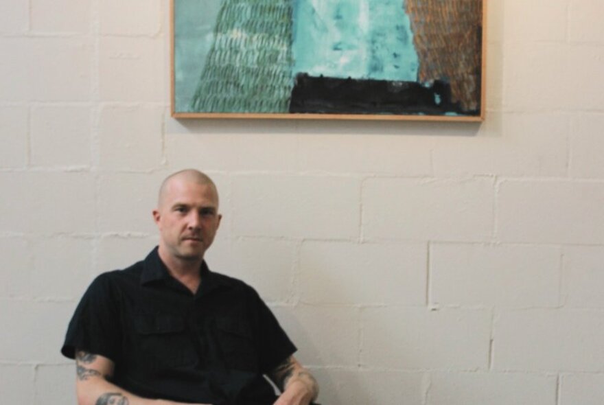 Artist Brendan Eccles seated in front of one of his paintings hanging on a white wall.