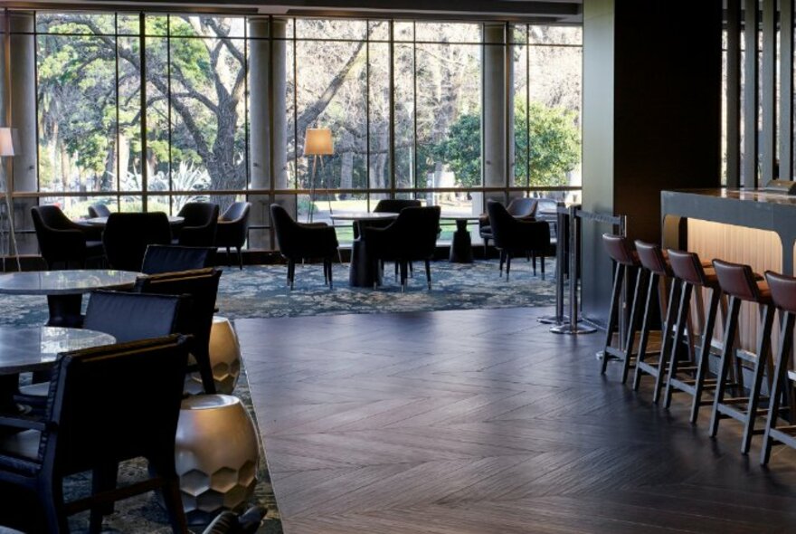 Interior of Cliveden restaurant showing parquetry floor and carpet, dining tables and chairs, a section of the bar and large floor to ceiling windows with views overlooking Fitzroy Gardens.